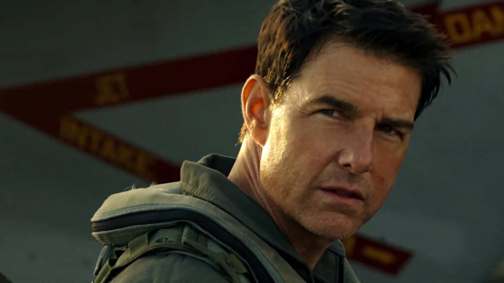Tom Cruise as Pete Mitchell in flight suit with military aircraft background Top Gun Maverick Wallpaper