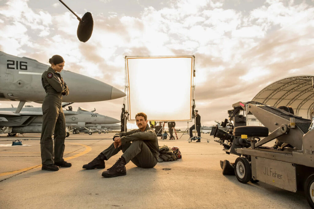 Top Gun Maverick cast in flight suits on airbase set with fighter jets and filming equipment Wallpaper