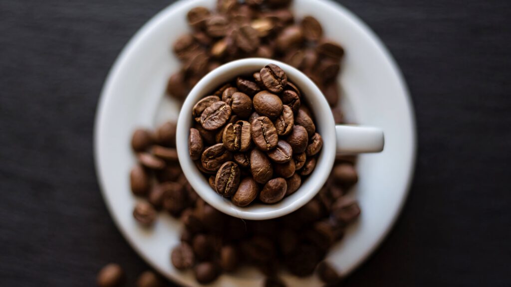 Top view of white cup with roasted coffee beans on saucer surrounded by beans on dark table Wallpaper
