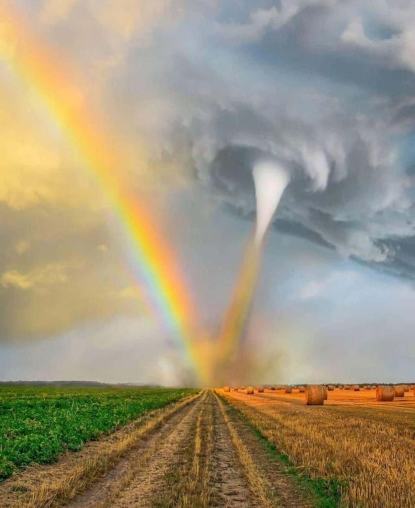 Tornado under vibrant rainbow over rural green crops golden hay bales dirt path landscape wallpaper