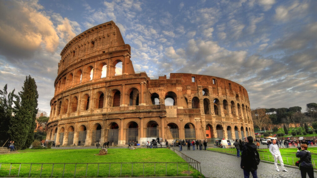 Tourists photographing the majestic Colosseum in Rome under a dramatic cloudy sky with green lawns Wallpaper