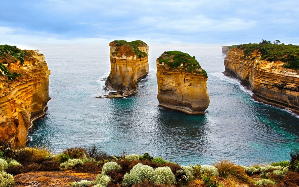 Towering coastal rocks rise from calm sea surrounded by lush greenery and distant island horizon Wallpaper