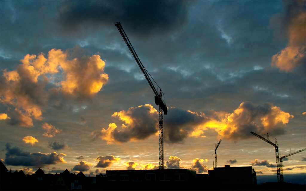 Silhouetted towering cranes at sunset with vibrant orange and blue clouds urban construction background Wallpaper