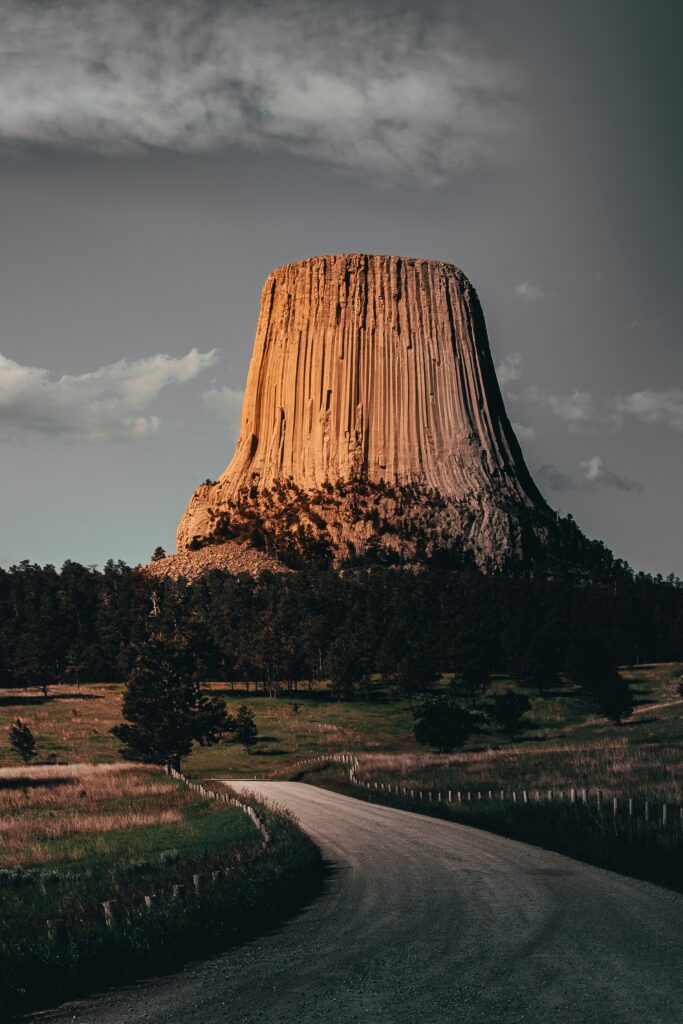 Towering rock formation above winding road surrounded by lush trees under soft sunlight sky Wallpaper