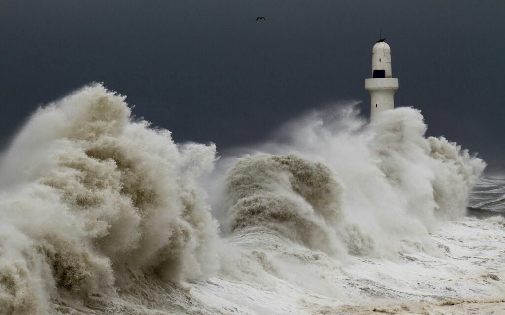Towering storm waves crash against white lighthouse under dark sky with soaring birds and sea foam Wallpaper