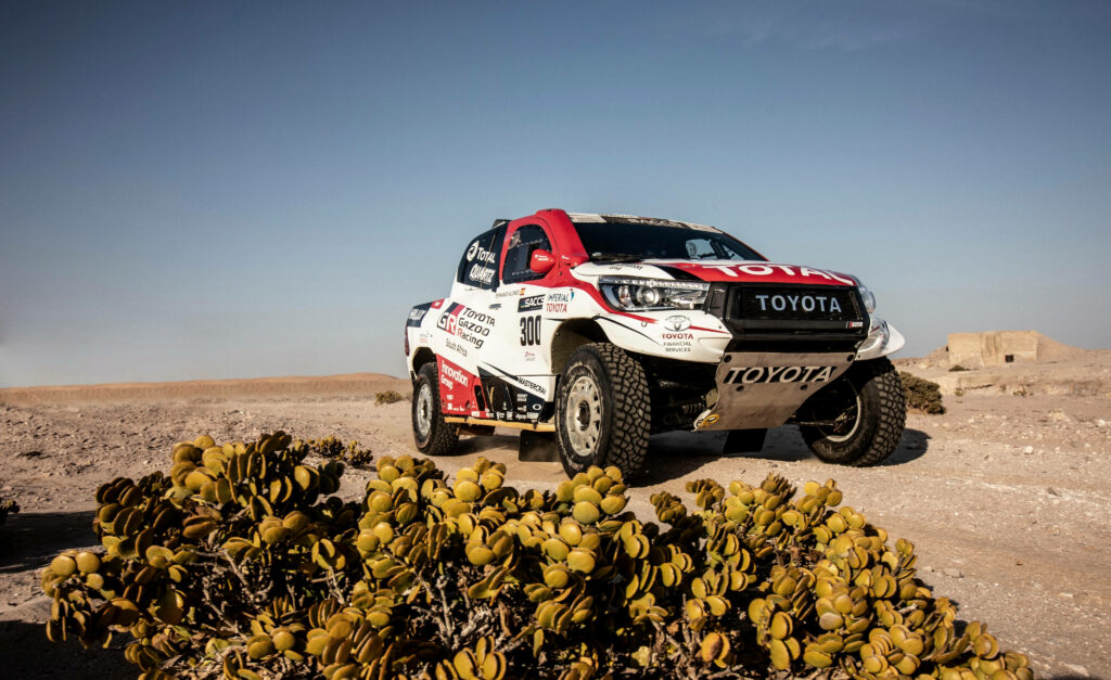 Red and white Toyota Hilux off-road vehicle parked on rugged desert terrain under clear sky Wallpaper