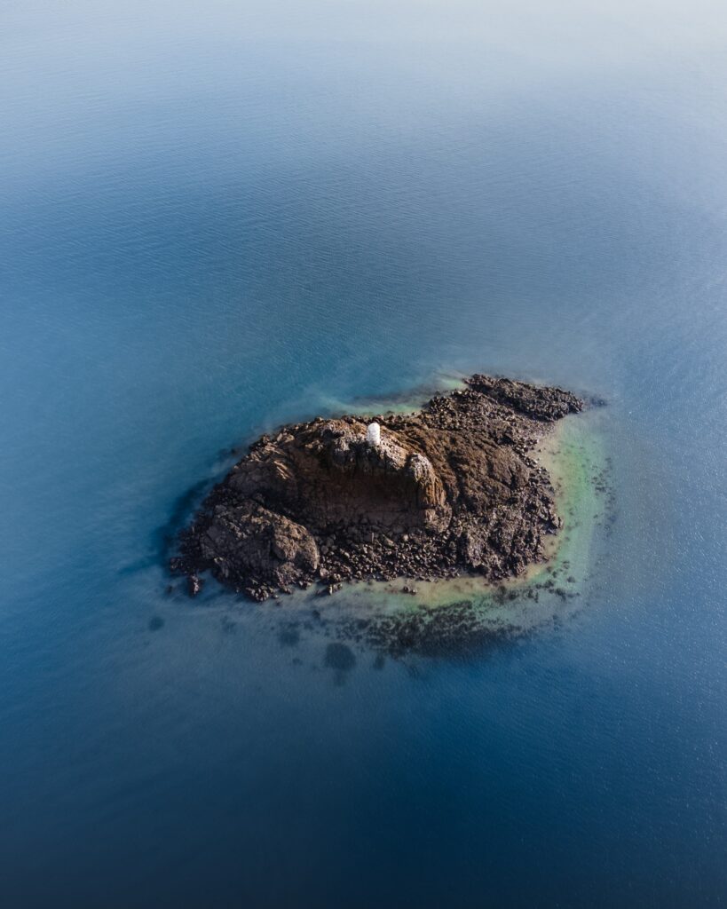 Aerial view of isolated island with lighthouse surrounded by calm blue sea and rocky shore Wallpaper