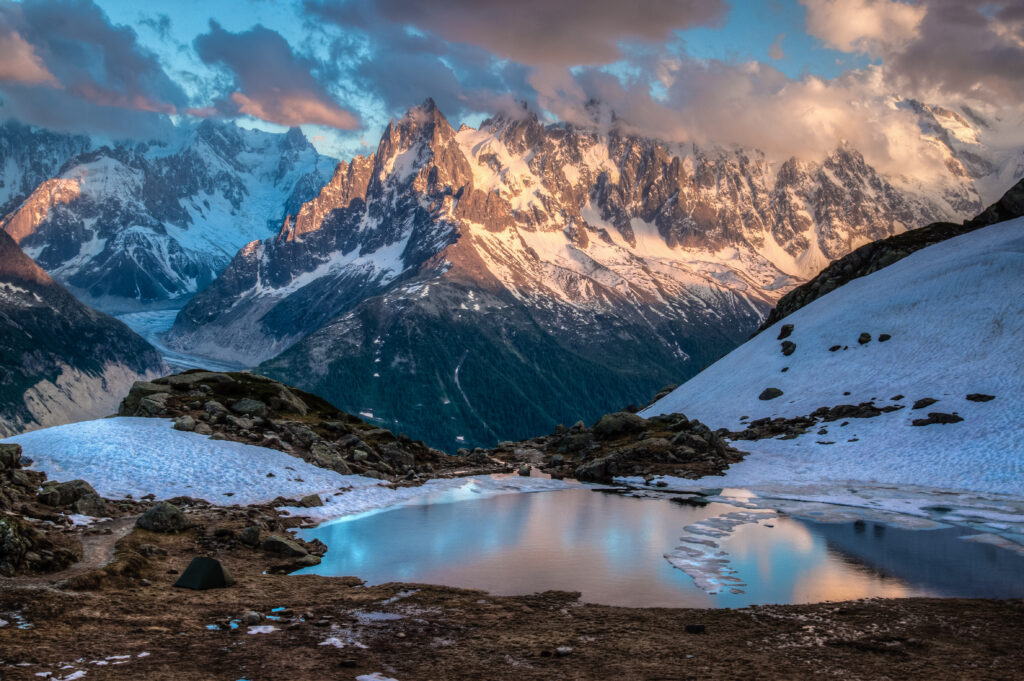 Tranquil alpine pond reflecting snow-covered mountains at sunset with melting snow patches Wallpaper