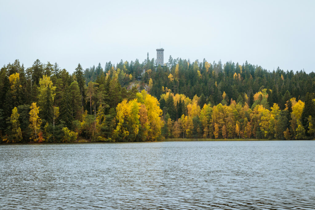 Serene lake reflecting autumn forest with evergreen trees and stone tower under overcast sky Wallpaper