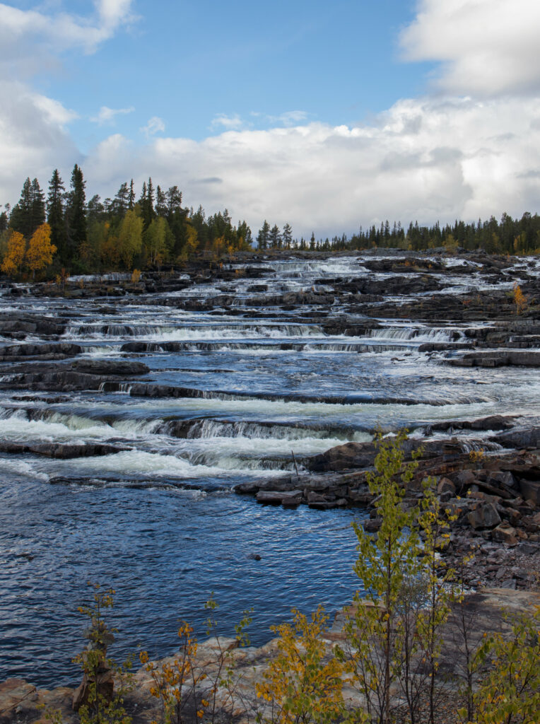 Peaceful river cascading over layered stones surrounded by autumn trees under cloudy sky wallpaper