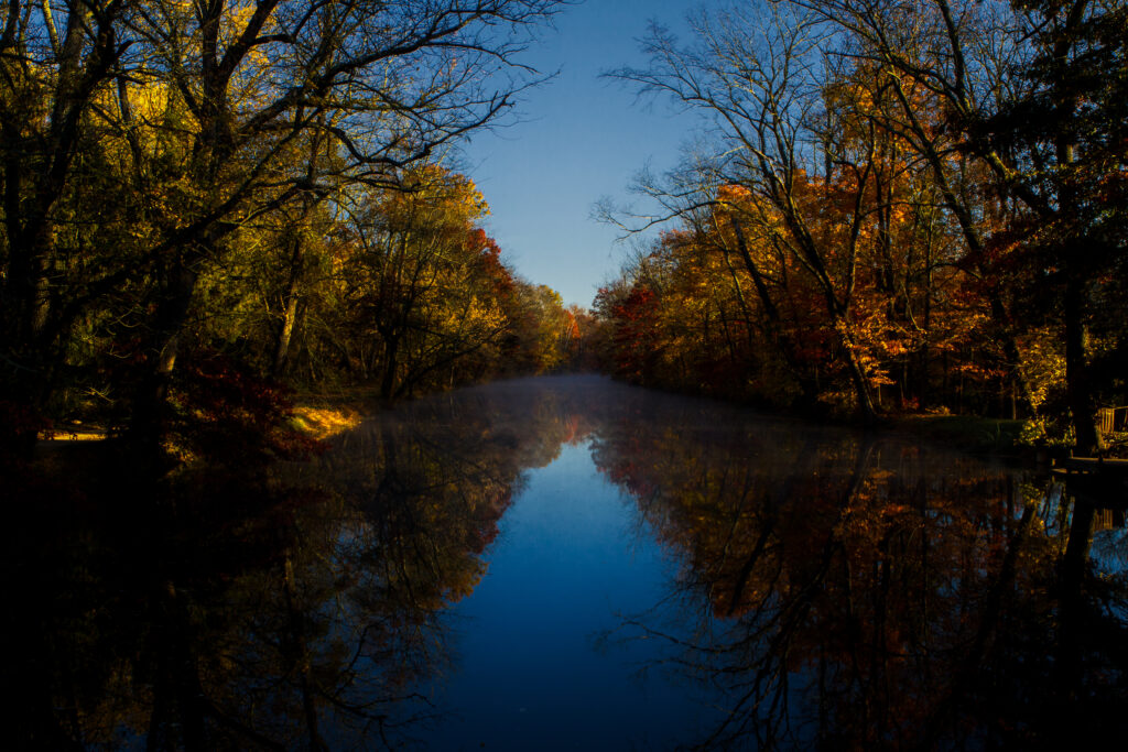 Serene autumn river with vibrant red and golden trees reflected in calm water under blue sky Wallpaper