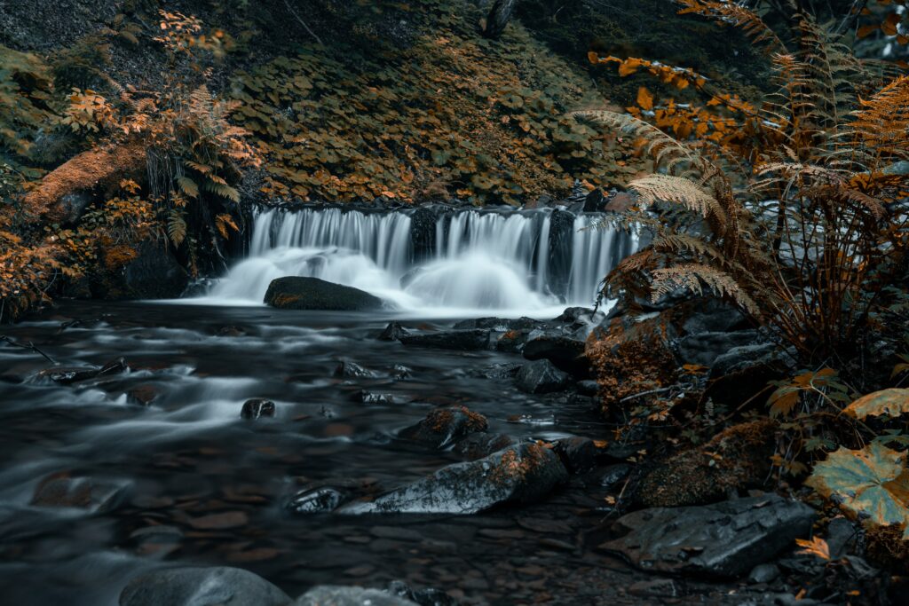 Serene waterfall flowing over rocks into stream with mossy stones and autumn foliage background Wallpaper