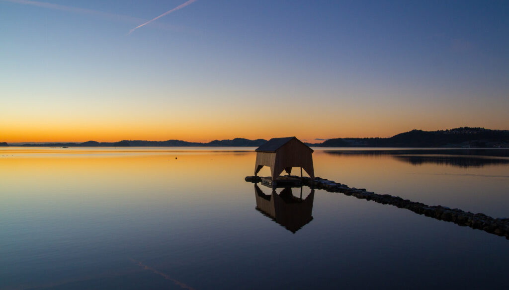 Tranquil beach house reflecting on calm lake at sunset with vibrant orange and blue sky colors Wallpaper