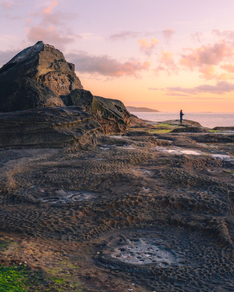 Tranquil sunset beach with rugged rocks and lone silhouette gazing over calm sea waters Wallpaper