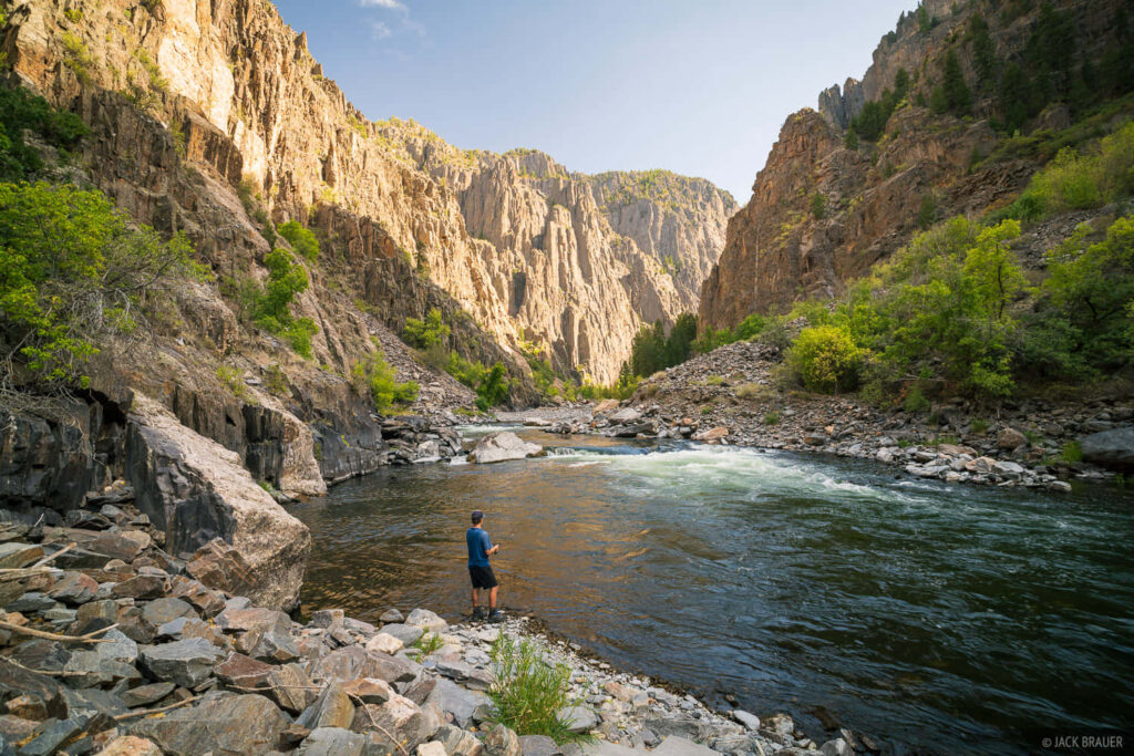 Person fishing on tranquil river beneath towering rugged cliffs in peaceful Black Canyon landscape Wallpaper