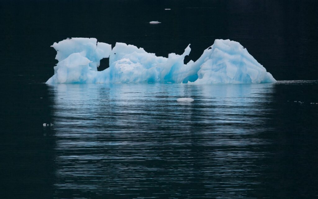 Serene blue iceberg floating on calm dark ocean with reflective water and distant mountains wallpaper