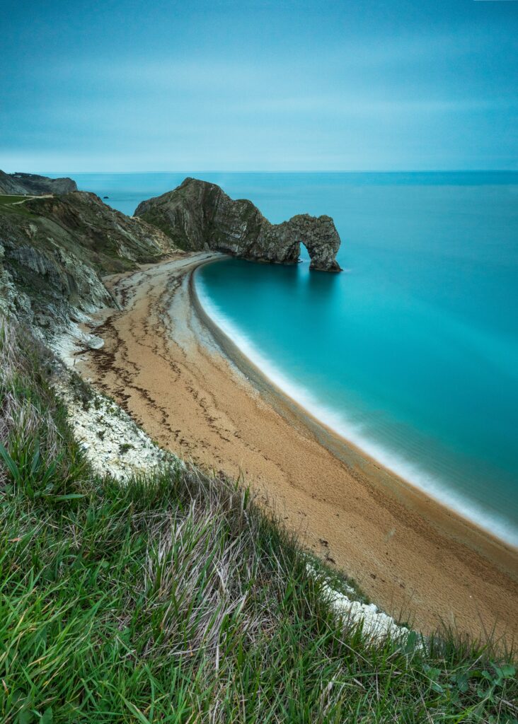 Tranquil coastal wallpaper with golden sand beach rugged rocks calm blue sea and grassy foreground