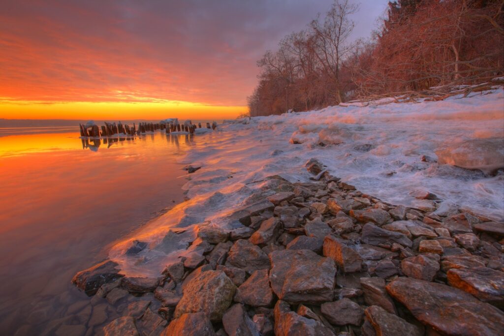 Tranquil coastal sunset with rugged stones gentle waves and leafless trees reflecting on water Wallpaper