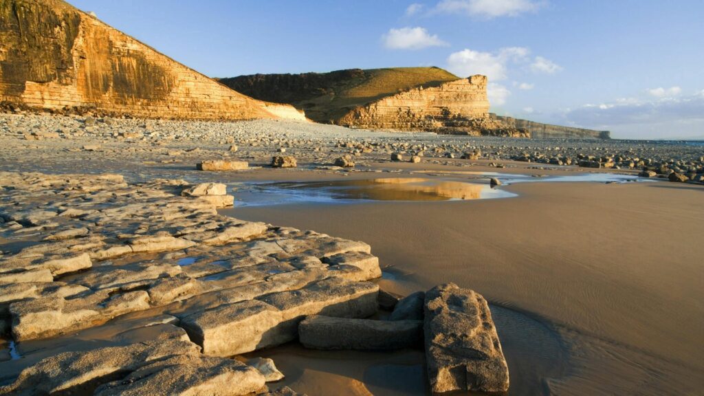 Scenic coastal landscape with weathered stones sand cliffs calm sea and golden sunlight background Wallpaper