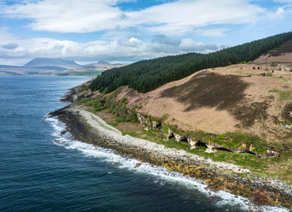Coastal landscape with rugged shore rolling hills evergreen forest and blue sea under clouds Wallpaper