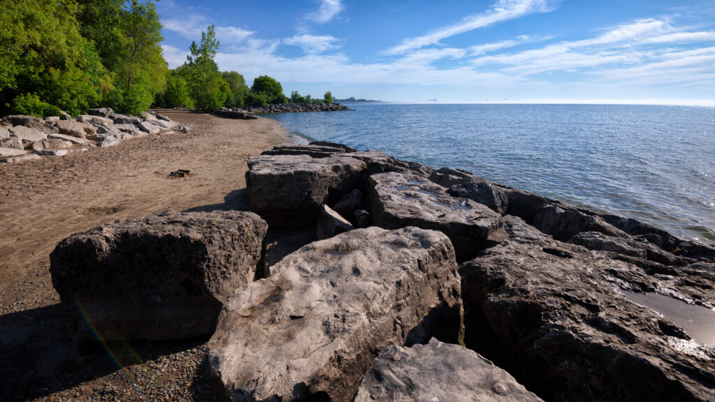 Tranquil coastal landscape with rugged stones sandy shore calm sea blue sky and green trees wallpaper