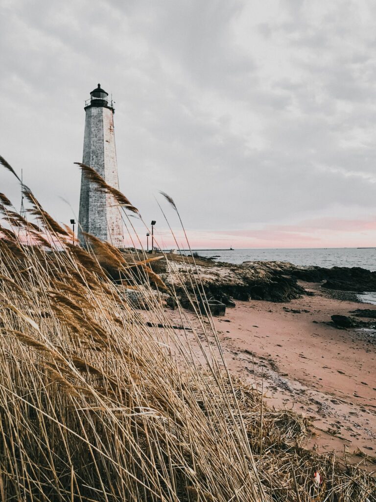 Tall weathered lighthouse on rugged coast with wild grass and calm sea at pastel sunset Wallpaper
