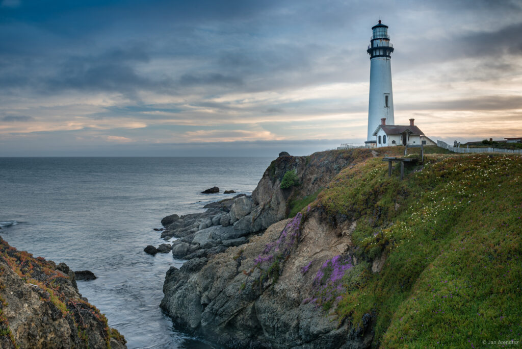 Lighthouse on rugged coastal cliffs with waves gentle sky and charming nearby building wallpaper background