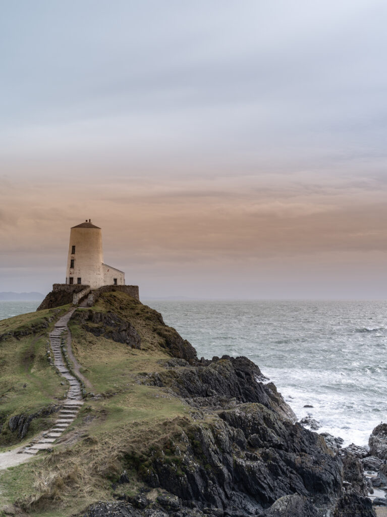 Solitary lighthouse on rocky cliffs with winding stone stairs overlooking restless sea at sunset Wallpaper