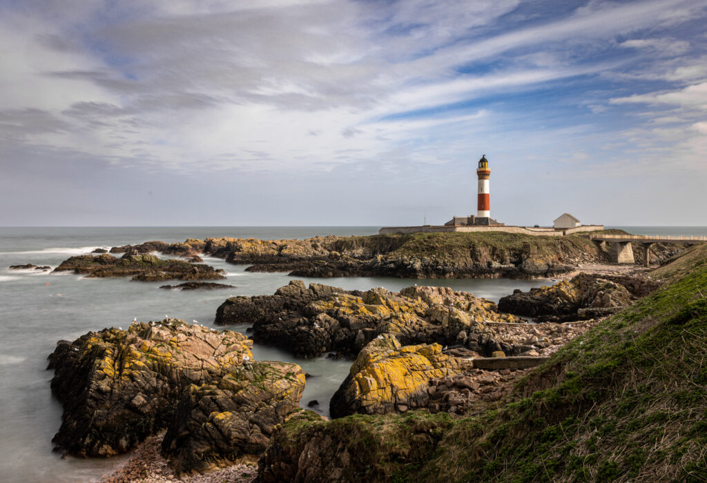 Lighthouse on rocky coast beside calm sea under partly cloudy sky with green grass background Wallpaper