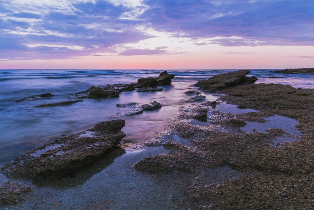 Serene coastal rocks extend into calm sea under pastel pink and purple sky for nature wallpaper