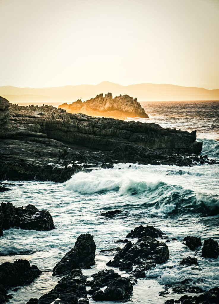 Rugged coastal rocks with dynamic waves gentle fog and warm sunlight on distant cliffs wallpaper