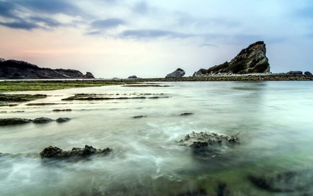 Tranquil coastal scene with gentle waves over rocky reefs and foggy pastel sky background Wallpaper