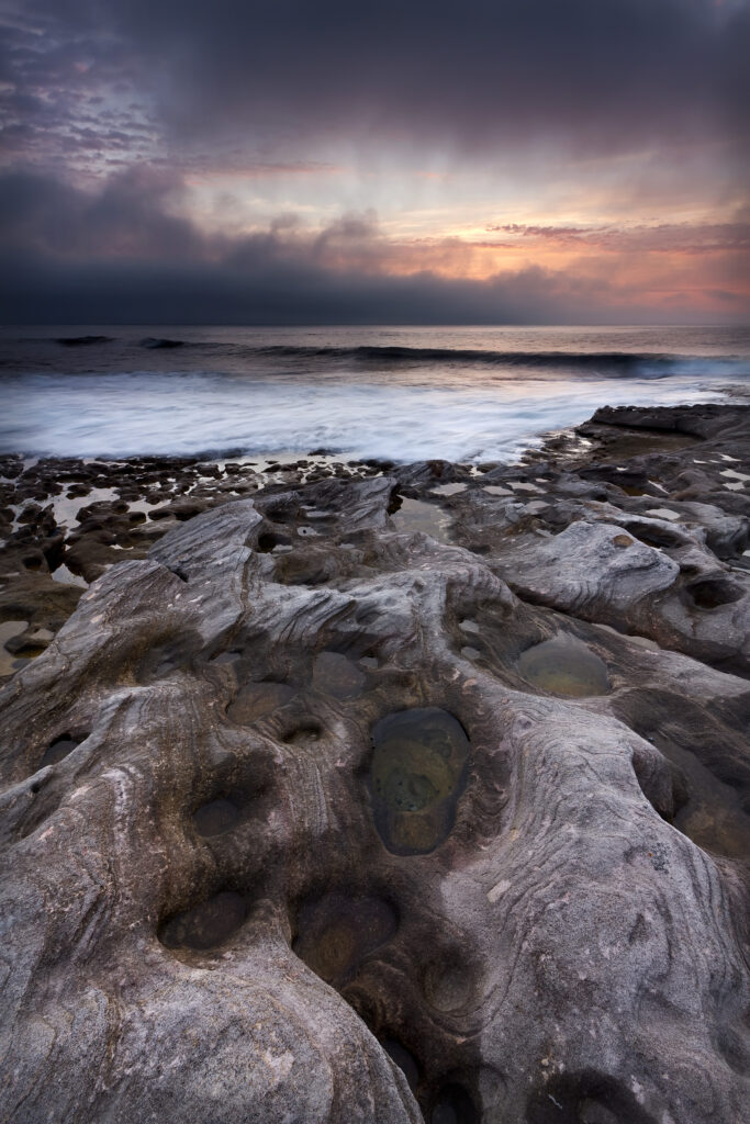 Rugged rocks by calm sea with gentle waves under vibrant orange purple blue sunset sky wallpaper