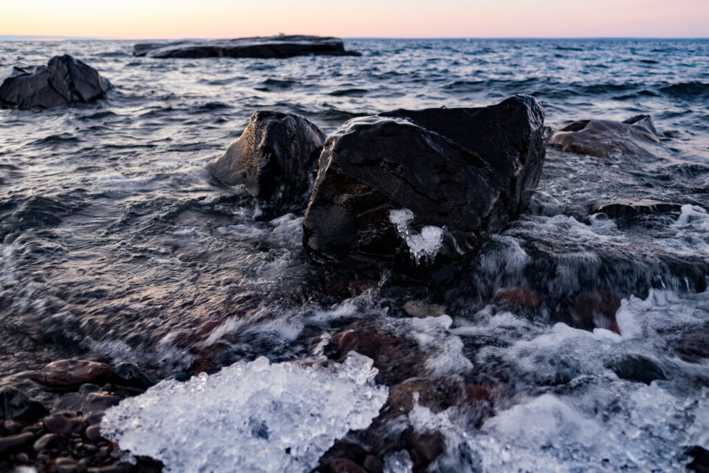Serene coastline with rugged stones icy shore and gentle waves under a warm twilight sunset Wallpaper