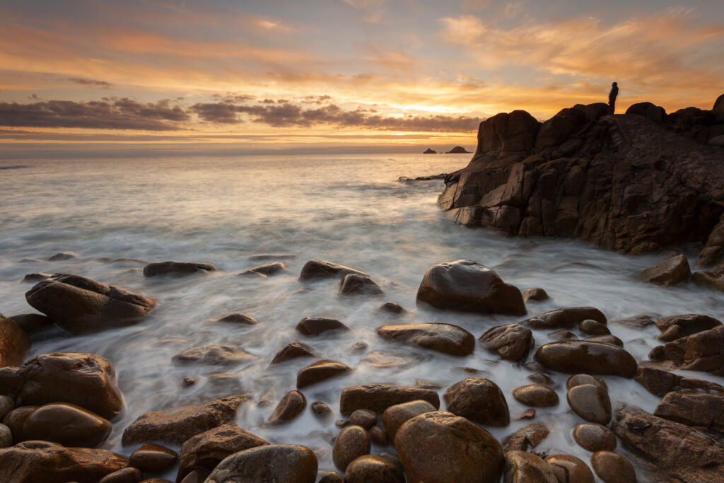 Serene coastal wallpaper with smooth rocks vibrant sunset waves and a lone figure standing