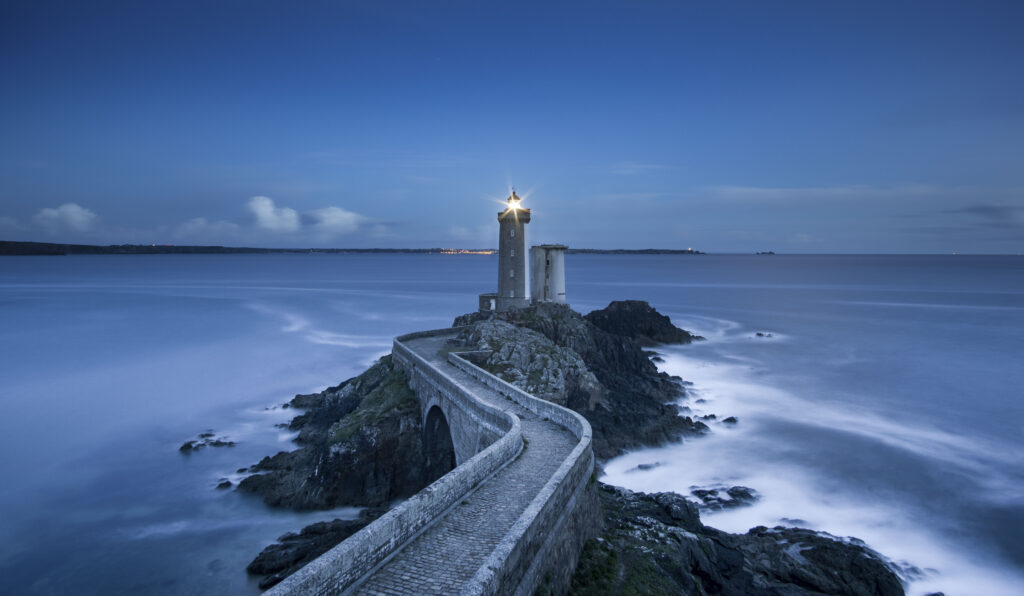 Tranquil coastal landscape with stone bridge and lighthouse over calm misty sea at twilight Wallpaper