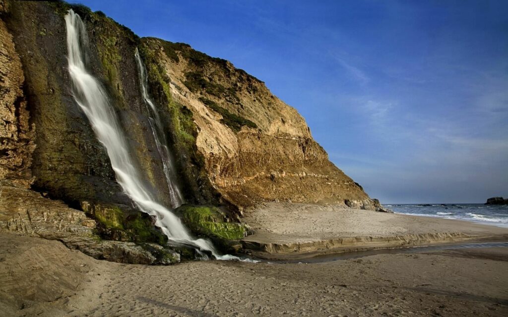 Coastal waterfall over mossy rocks onto golden sand under vivid blue sky natural landscape wallpaper