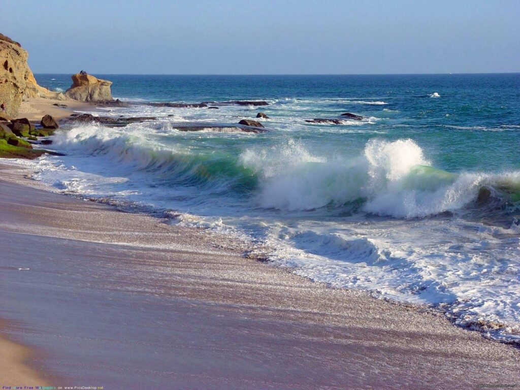 Coastal wallpaper with turquoise waves crashing on sandy shore and rugged rocks under clear sky