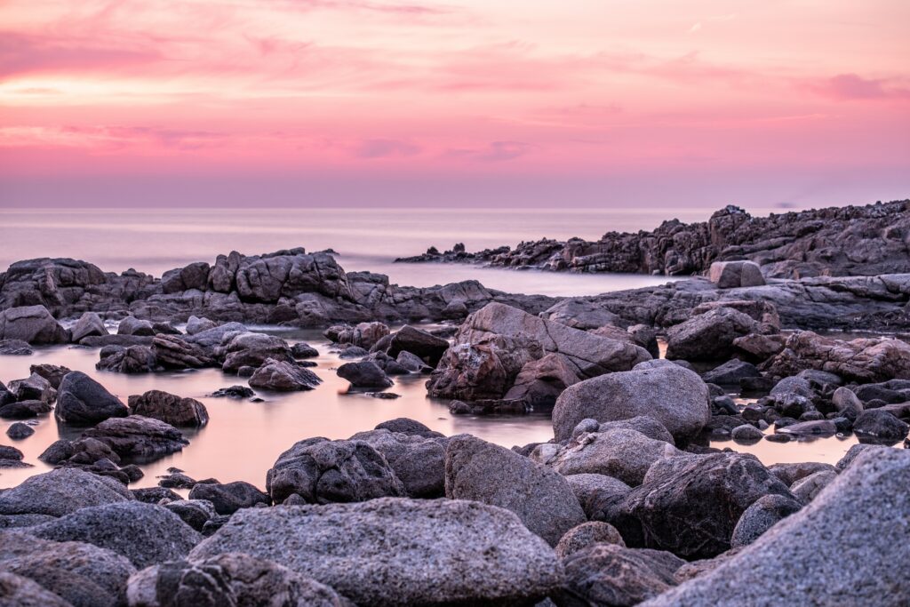 Tranquil coastline at dusk with rugged rocks smooth stones and pastel pink purple sky reflection Wallpaper
