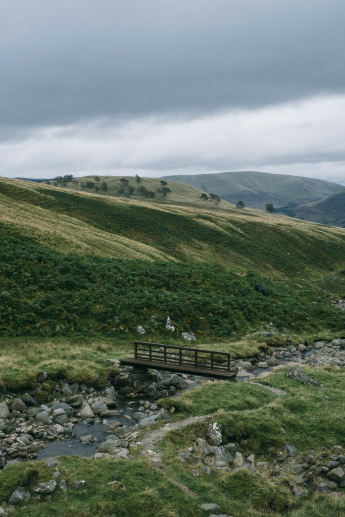 Rolling green hills with wooden bridge over clear stream serene countryside wallpaper background