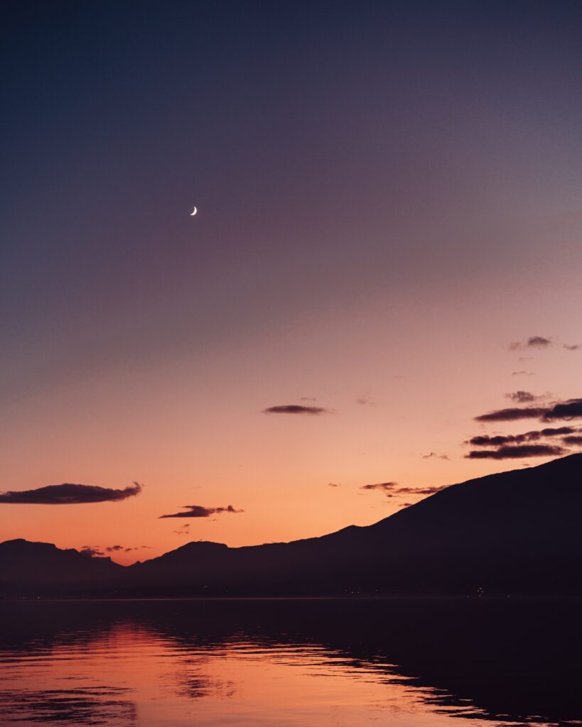 Crescent moon above purple orange dusk sky reflects on calm lake with silhouetted mountains wallpaper