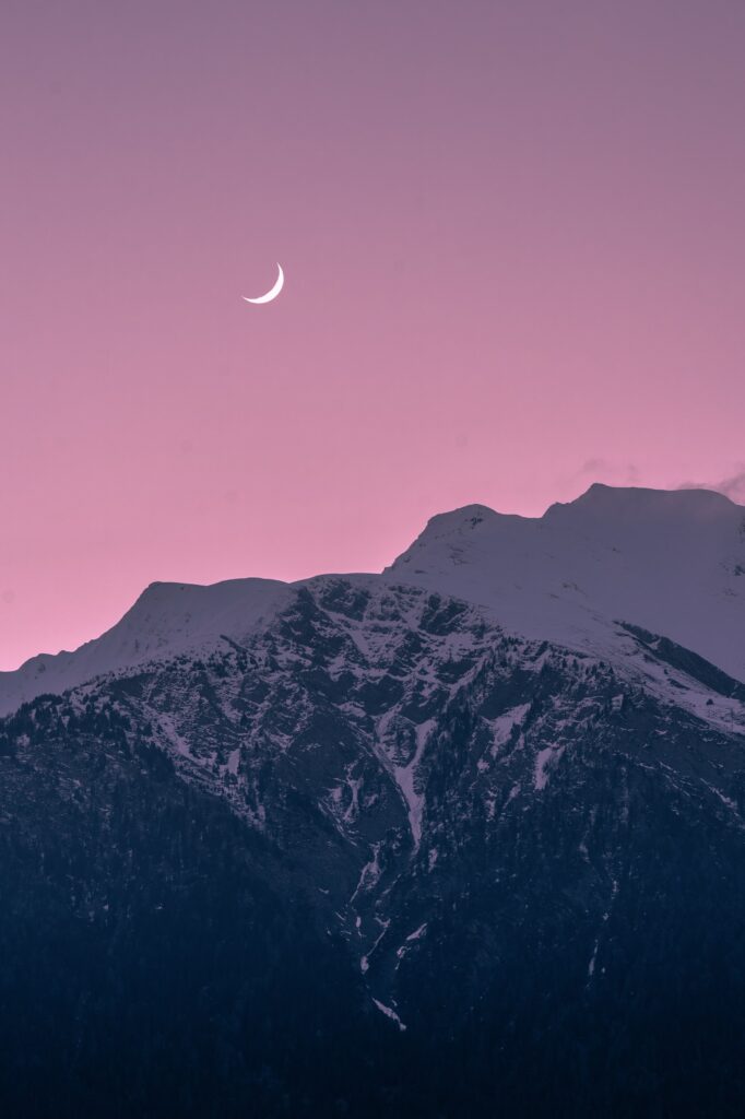 Crescent moon over snow-capped mountains and rocky slopes at pink purple winter twilight wallpaper