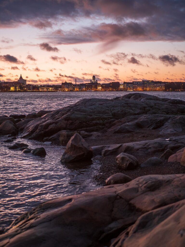 Rugged rocks by calm sea with city skyline and glowing buildings under dusk sky wallpaper background