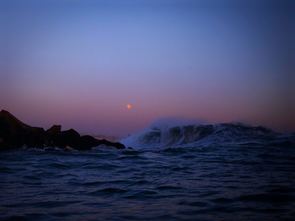 Tranquil dusk seascape with deep blue waves crashing rugged rocks under pastel sky and full moon Wallpaper