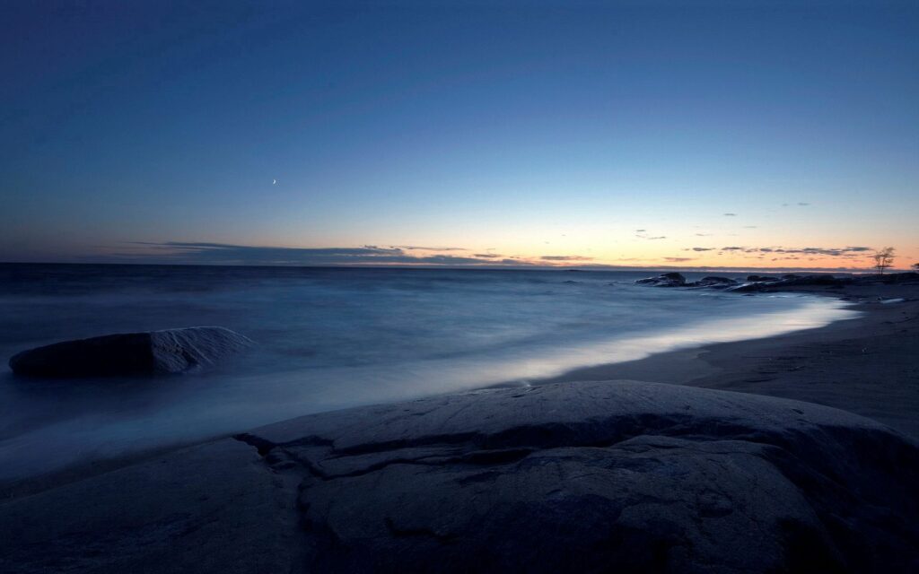 Tranquil dusk shore with soft waves over smooth rocks and glowing blue horizon wallpaper background