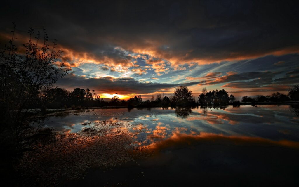 Evening sky with fiery sunset clouds reflecting on calm lake surrounded by dark tree silhouettes Wallpaper