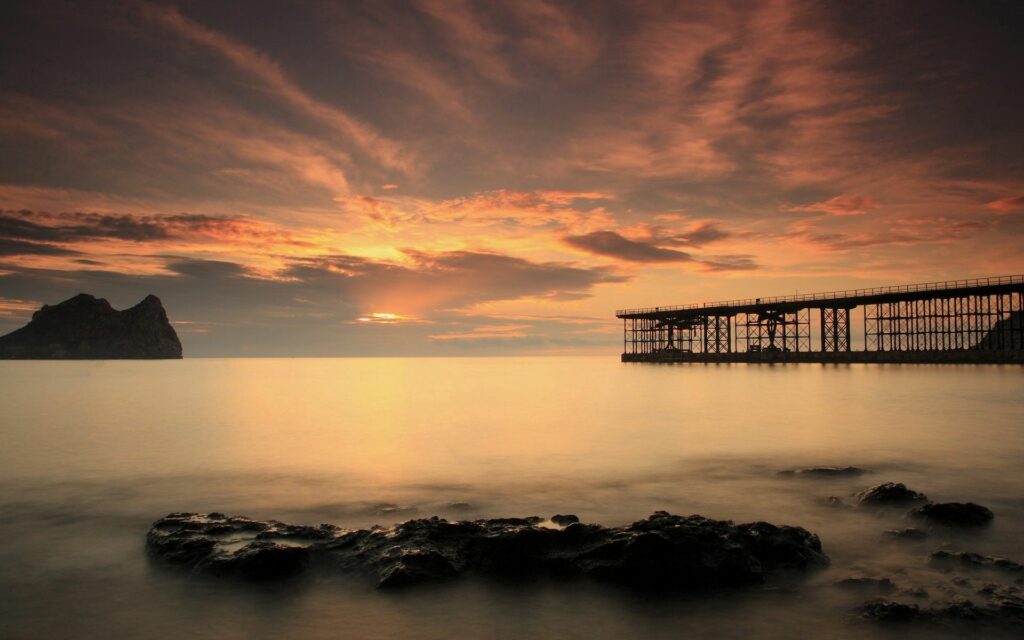 Tranquil evening sky with warm orange hues over rustic bridge and calm water landscape wallpaper