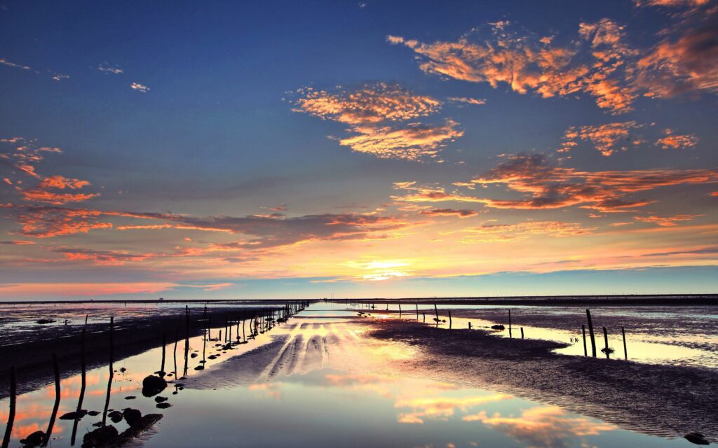 Sunset over sandy shoreline with stakes reflected in shallow wetland pools and colorful sky Wallpaper