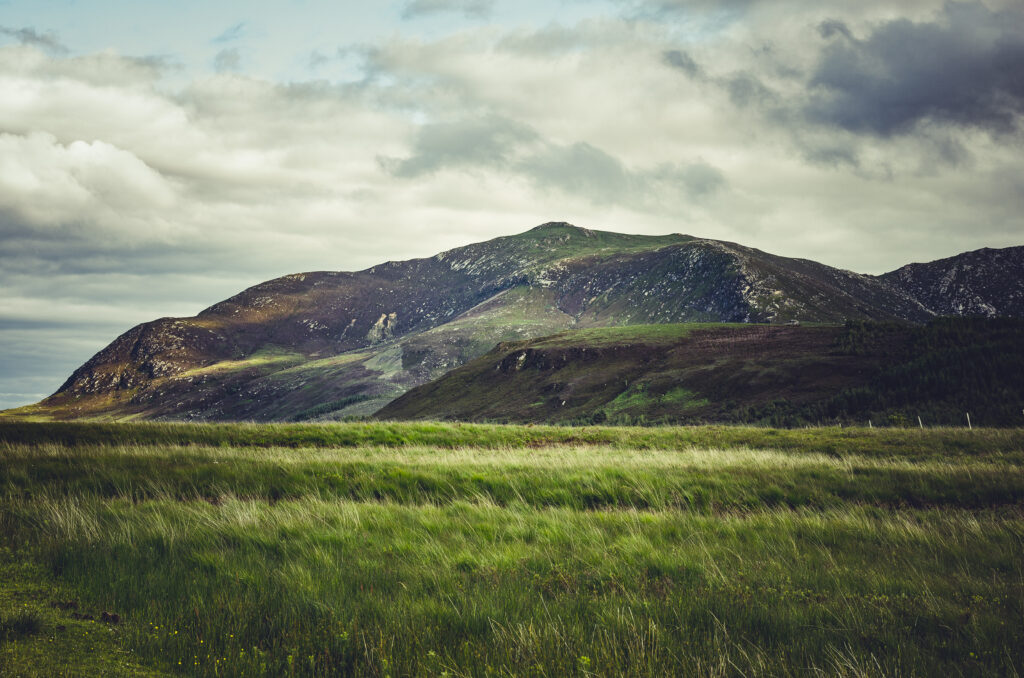 Serene landscape with rolling green hills grassy valley rugged mountains and cloudy sky wallpaper
