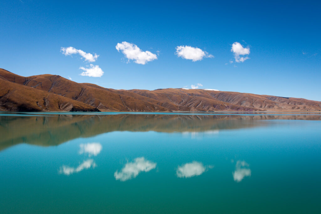 Tranquil earthy hills reflect on serene lake under blue sky with fluffy white clouds wallpaper background